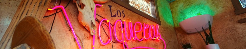 A vibrant Mexican-themed interior features a neon sign reading "Los Vaqueros" beside a bull skull and antlers. Warm lighting creates a cozy ambiance.