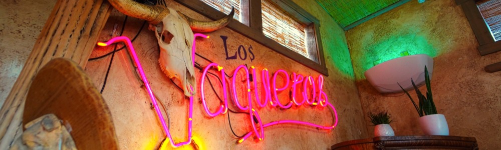 A vibrant Mexican-themed interior features a neon sign reading "Los Vaqueros" beside a bull skull and antlers. Warm lighting creates a cozy ambiance.